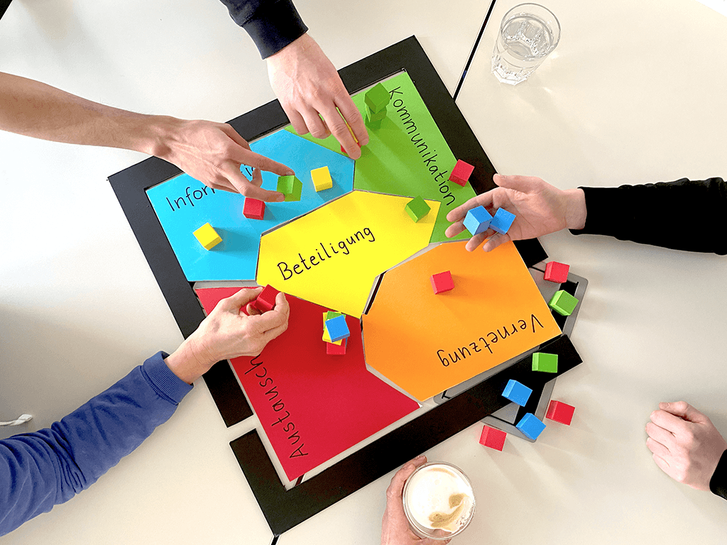 Hände legen bunte Bausteine auf ein Beteiligungsspiel mit den Feldern Information, Kommunikation, Beteiligung, Vernetzung und Austausch